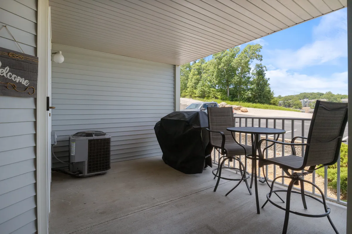 A welcoming exterior scene showing an outdoor seating area complete with a grill and a prominent welcome sign. The space is designed for comfortable outdoor dining, with chairs and a table set beneath a screened-in or covered patio area. The backdrop features sunset views, providing a warm, inviting atmosphere for communal gatherings and meals.
