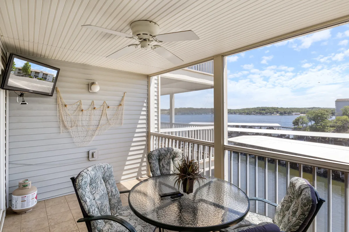A glass-topped table with comfortable seating is set against a backdrop of wide water views, all within a screened enclosure. Decorative netting adds a nautical touch, and a wall-mounted TV suggests comfort and entertainment for longer stays on the deck.