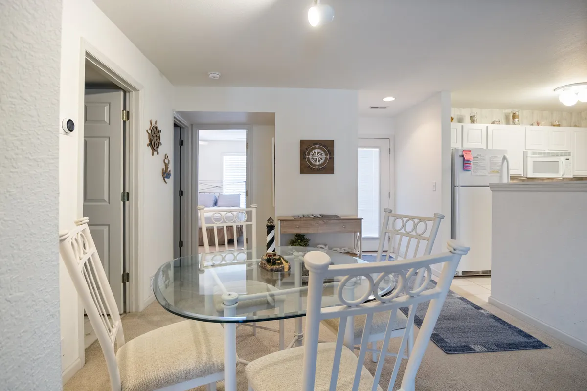 A cozy dining area is depicted, with a glass dining table surrounded by white chairs. Decorative wall art and a glimpse of the kitchen area in the background create a seamless transition between cooking and dining spaces.