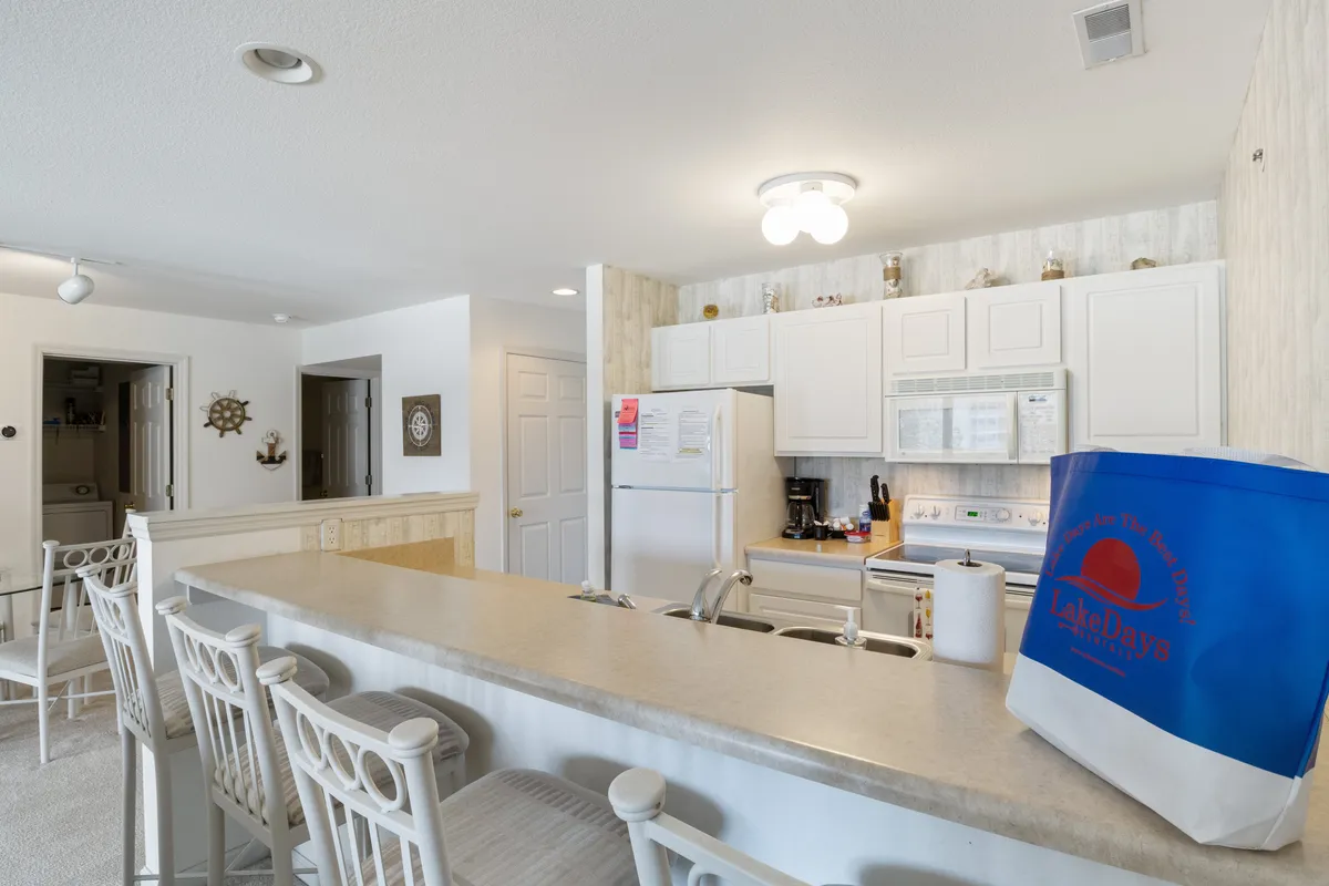 This image features a bright kitchen with white cabinetry, a kitchen island with seating, and a variety of decorative items on the shelves. Modern appliances and inviting bar stools make the space feel both functional and social, ideal for preparing meals and casual dining.