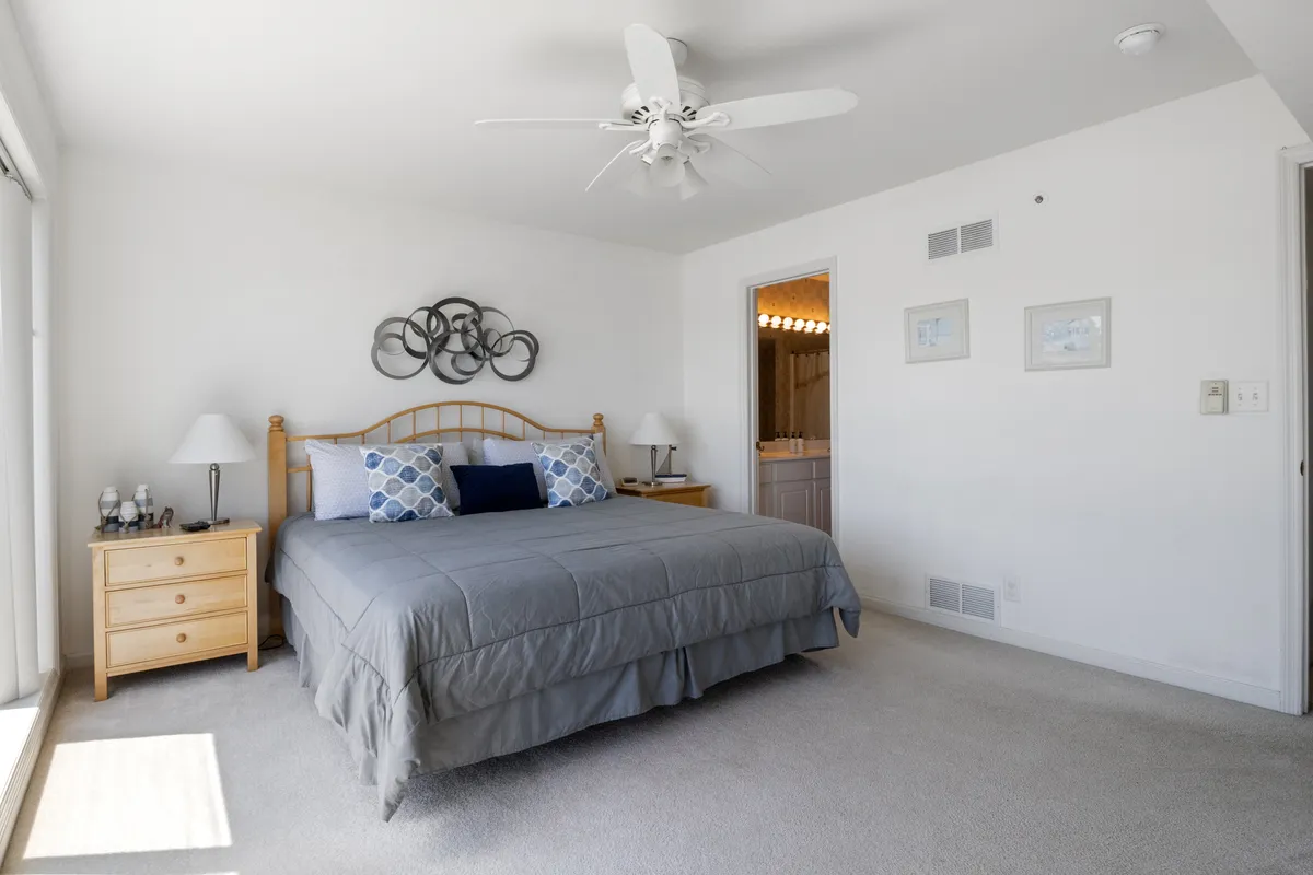 A tranquil bedroom with a comfortable bed dressed in gray bedding and blue accent pillows. A nightstand with a softly lit lamp and decorative wall art enhance the peaceful atmosphere, while sunlight streams in, illuminating the space and suggesting inviting mornings.