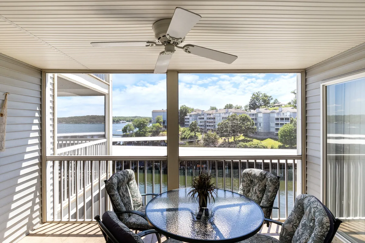 This image features a screened-in deck with a dining table with a glass top and green patterned chairs. The ceiling fan above and the panoramic lake view beyond the screen create a tranquil, airy retreat. The setup is perfect for outdoor dining or relaxing with a book while enjoying the scenery.