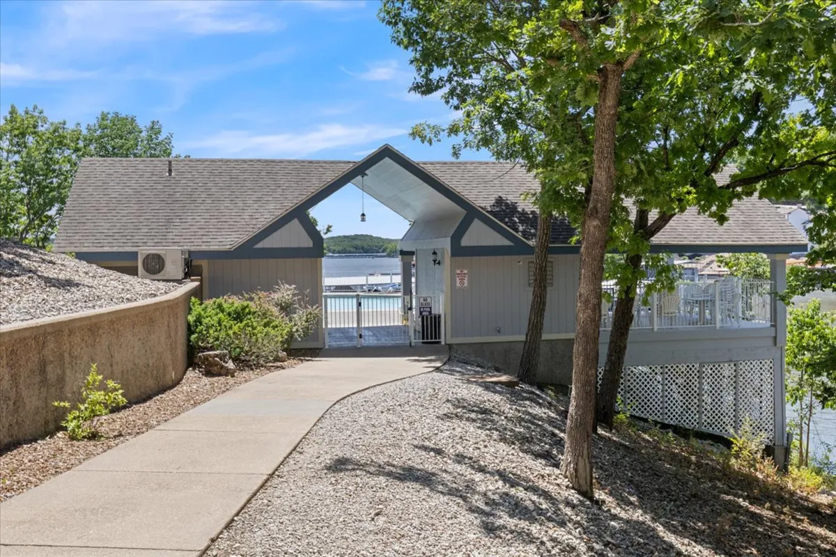 An exterior shot of the building's entrance framed by a scenic pathway, lush landscaping, and mature trees, with a glimpse of the water in the background. The composition evokes a sense of arrival, tranquility, and proximity to nature, suggesting easy access to lakeside activities and serene surroundings.