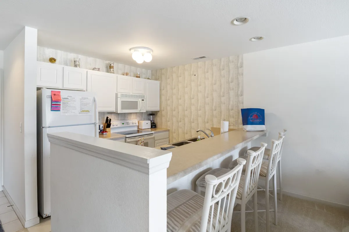 The image shows a welcoming kitchen with a central island, bar stools for casual seating, and modern appliances arranged neatly along the walls. The space is styled for both cooking and socializing, with ample countertop space and visible cookware ready for use.