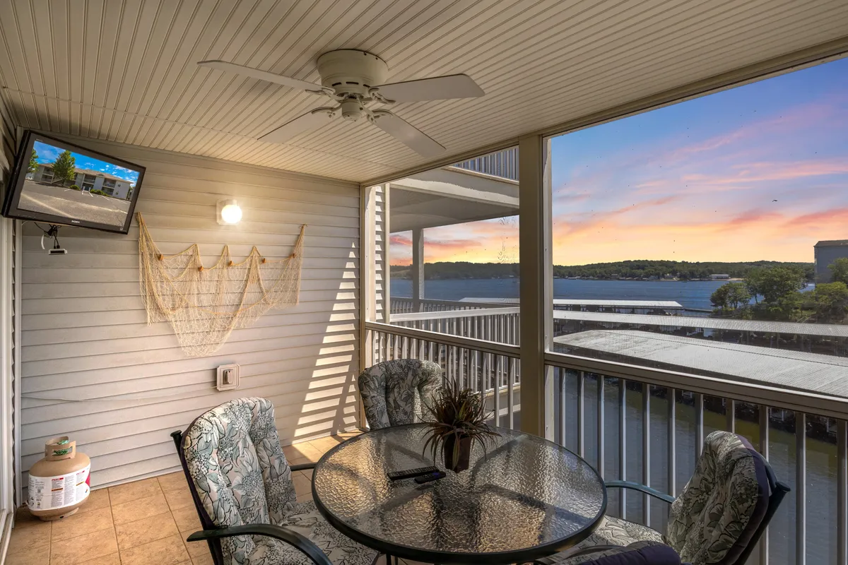 The image showcases a serene, screened-in deck overlooking Lake Ozark’s main channel at sunset. The space features a glass-top table surrounded by comfortable chairs, perfect for relaxing or sharing a meal. Decorative wall art adds a touch of personality, while large windows connect the indoor and outdoor experience. The view captures the tranquil water bathed in golden sunset light, inviting guests to unwind and soak in the scenery.