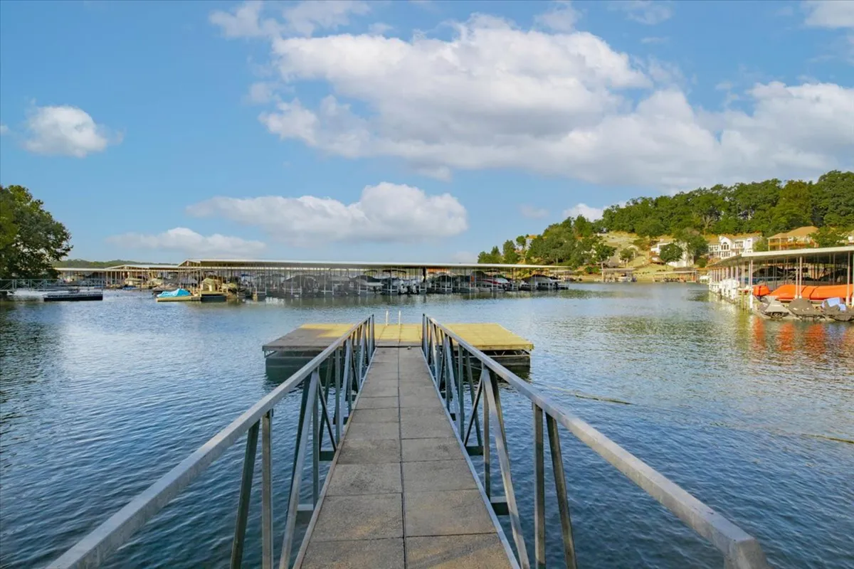 A tranquil lakeside dock stretches out over calm water, with boats moored alongside and gentle hills visible in the background under a wide sky. The scene is peaceful, inviting guests to imagine boating, fishing, and water-based fun steps from the condo.