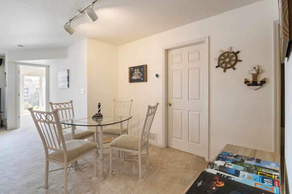 This bright, welcoming dining area features a glass table with four white chairs and nautical-themed decor. Art on the walls and ample natural light create a cheerful ambiance, making it an inviting spot for shared meals, conversations, or board games.