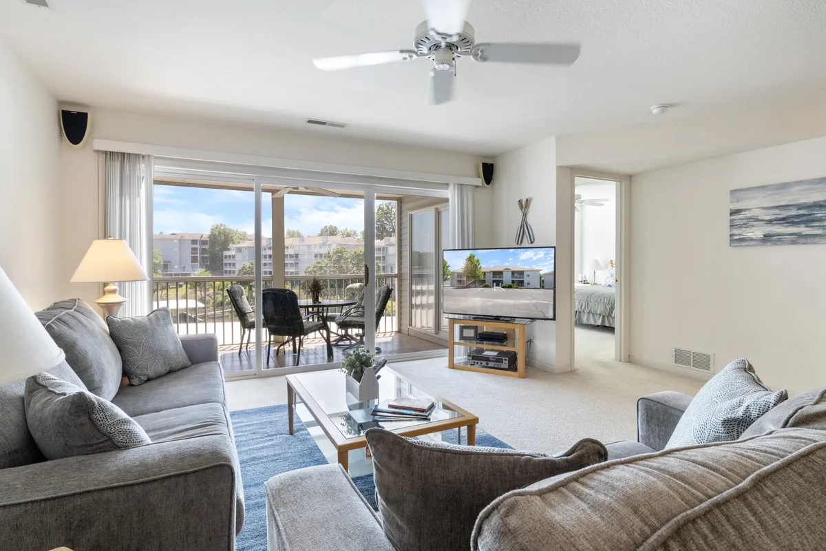 This image depicts a cozy, inviting living area featuring comfortable gray sofas, a sleek glass coffee table, a large modern television, and expansive windows offering panoramic views of the lake. The space is bathed in natural light, with plush seating arranged to encourage conversation and relaxation.